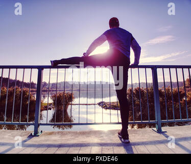 Mann stretching Beine, bevor sie nach draußen gehen in sonniger Morgen laufen. Aufwärmen der Muskeln. Im freien Training auf See brücke. Stockfoto