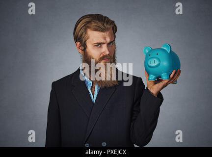 Finanzen, Geld. Ernster Mann hält Sparschwein. Der bärtige Mann hält piggy Bank voll Geld über grauen Hintergrund. Geschäftliche und Finanzielle; Finanzen messen Stockfoto