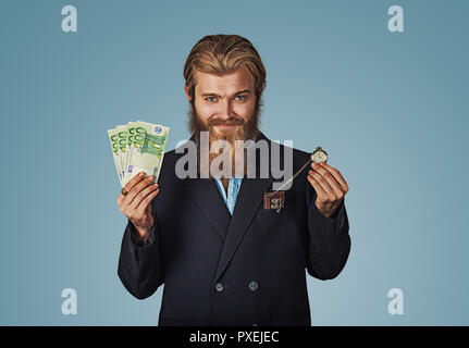 Zeit ist Geld. Schöne fröhliche bärtiger Hippie Mann lächelnd mit Bargeld und Taschenuhr. Lustiger Kerl ist ein glücklicher Gewinner Holding ein Haufen Geld und ein Wat Stockfoto