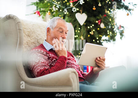 Einen überraschten älteren Mann sitzt auf einem Sessel durch einen Weihnachtsbaum, mit Tablette. Festliche Zeit. Stockfoto