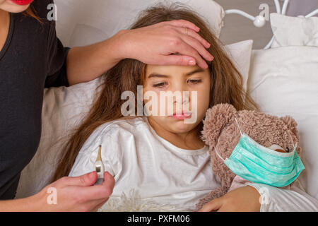 Ein kleines Mädchen mit ihrem Liebling Teddybär auf dem Sie trägt eine Mullbinde ist krank im Bett und ihre Mutter misst sie Temperatur mit einer Elek. Stockfoto