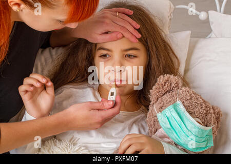 Ein kleines Mädchen mit ihrem Liebling Teddybär auf dem Sie trägt eine Mullbinde ist krank im Bett und ihre Mutter misst sie Temperatur mit einer Elek. Stockfoto