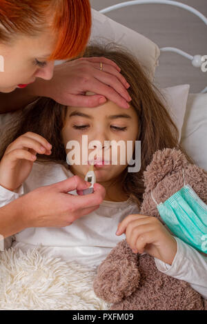 Ein kleines Mädchen mit ihrem Liebling Teddybär auf dem Sie trägt eine Mullbinde ist krank im Bett und ihre Mutter misst sie Temperatur mit einer Elek. Stockfoto