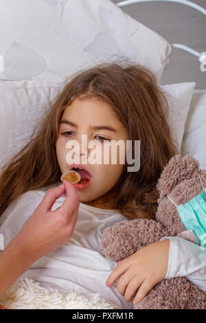 Ein kleines Mädchen mit einem bevorzugten Teddybär auf denen Sie auf eine Mullbinde, liegt krank im Bett und ihre Mutter trinkt ihr Medizin. Stockfoto