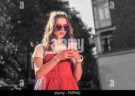 Mädchen mit roten Gläsern und Kleid Holding ihr rosa Smartphone sms-Freund Stockfoto