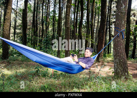 Happy asiatischer Mann mit Smartphone liegen in der Hängematte auf der Wald Stockfoto