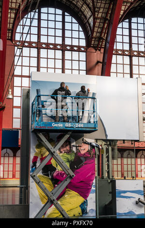 Antwerpen, Belgien - 2018-10-01: Hoch über einem belebten Bahnhofshalle, auf einem highworker Gerüst Plattform auf Rädern, zwei Männer beschäftigt sind, einen riesigen advertisem Stockfoto