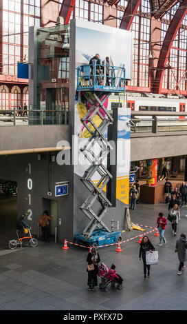 Antwerpen, Belgien - 2018-10-01: Hoch über einem belebten Bahnhofshalle, auf einem highworker Gerüst Plattform auf Rädern, zwei Männer beschäftigt sind, einen riesigen advertisem Stockfoto