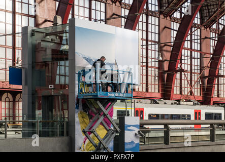 Antwerpen, Belgien - 2018-10-01: Hoch über einem belebten Bahnhofshalle, auf einem highworker Gerüst Plattform auf Rädern, zwei Männer beschäftigt sind, einen riesigen advertisem Stockfoto