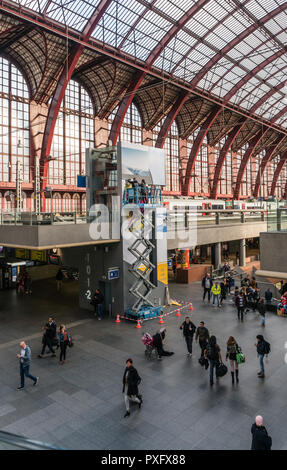 Antwerpen, Belgien - 2018-10-01: Hoch über einem belebten Bahnhofshalle, auf einem highworker Gerüst Plattform auf Rädern, zwei Männer beschäftigt sind, ein riesiges werben Stockfoto