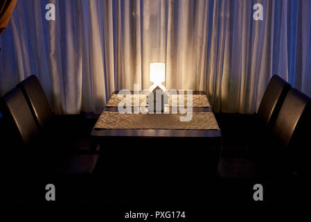 Brennende Lampe auf dem Tisch in der Nähe der Fenster Vorhang in der Dämmerung. Stockfoto
