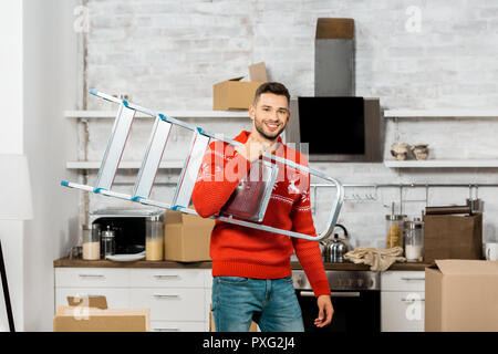 Fröhlicher junger Mann, der Leiter in Küche mit Kartons beim Umzug in neues Zuhause Stockfoto