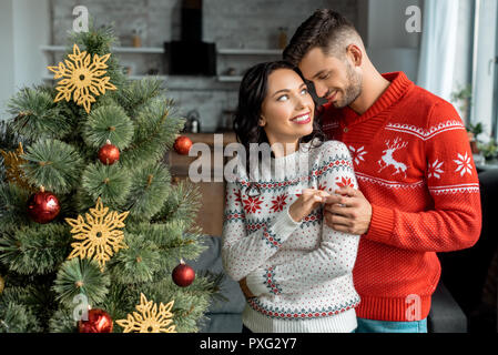 Lächelnde junge Paar umarmen und an jedem anderen suchen in der Nähe von Weihnachten zu Hause Stockfoto