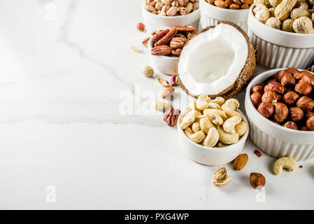 Verschiedene Arten von Nüssen - Walnüsse, Pekannüsse, Erdnüsse, Haselnüsse, Mandeln, Kokos, Cashews, in Schalen, auf einem weißen Marmor Tabelle Ansicht von oben Stockfoto