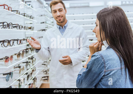 Selektiver Fokus der männlichen Augenarzt, der von Hand in Regalen mit Brillen zu nachdenkliche Frau, die in der Augenheilkunde shop Stockfoto