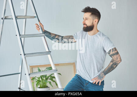 Junger bärtiger Mann mit Tätowierungen an Leiter im neuen Haus Stockfoto
