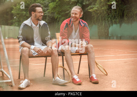 Gerne sportliche Freunde mit hölzernen Schläger sitzen auf Stühlen am Tennisplatz Stockfoto