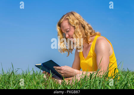 Junge kaukasier Frau liest Buch im Gras mit blauem Himmel Stockfoto