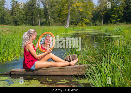 Blonde junge niederländische Frau sitzt mit Spiegel am Wasser in der Natur Stockfoto