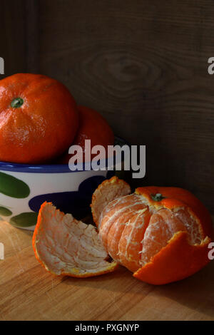 Helles orange Clementinen in blauen und weißen Schale und eine teilweise geschält Clementine auf Holzbrett mit dunklen Hintergrund Stockfoto