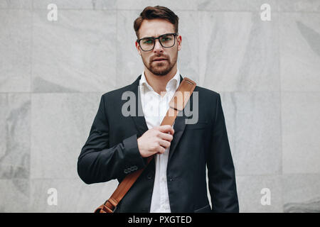 Geschäftsmann, gegen einen gefliesten Wand tragen ein Büro Tasche. Junger Mann Brillen tragen und Durchführung einer Office bag Stand im Freien. Stockfoto