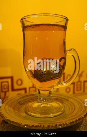 Heißen Kamillentee in transparenten Tee Tasse mit niedlichen Tea Leaf Filter vor der gelben wand serviert. Stockfoto