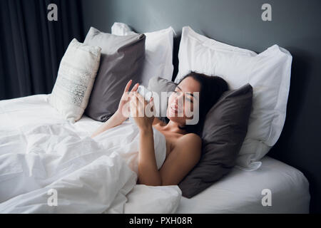 Brunette Mädchen liest Sms von ihrem Freund auf mobilen, während liegt im Bett. Fröhliche junge Frau von Decke bedeckt ist die Wiedergabe von Videos auf Handy vor und nach dem Schlafen. Stockfoto