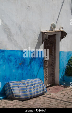 An der Wand eines Hauses mit einem Teil durch helle blaue Farbe lackiert. Reich verzierte Tor. Eine alte Matratze neben der Wand. Gepflasterte Gasse mit Kopfsteinpflaster. Kasbah de Oudaias, Stockfoto