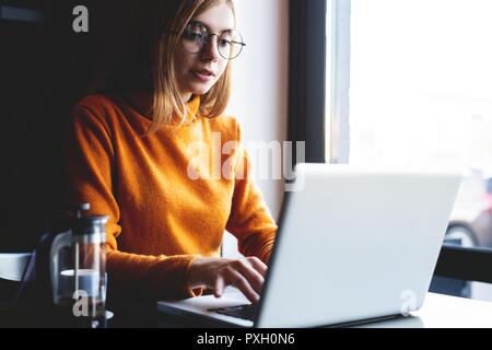 Bei der Arbeit konzentriert. Selbstbewussten jungen Frau in Smart Casual Wear Arbeiten am Laptop, während in der Nähe der Fenster in kreative Büro oder Cafe sitzen. Stockfoto