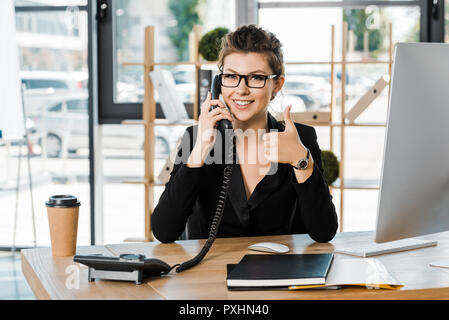 Lächelnd attraktive Geschäftsfrau sprechen durch stationäre Telefon im Büro und mit Daumen hoch Stockfoto