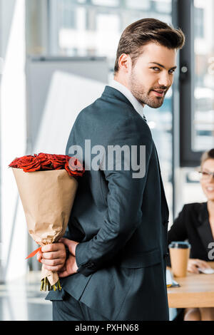Lächelnd gut aussehender Geschäftsmann ausblenden Blumenstrauß aus Rosen hinter zurück Geschäftsfrau im Amt zu lassen sie sich überraschen Stockfoto