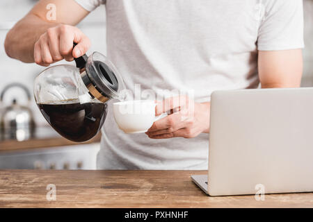 Zugeschnittenes Bild des Menschen Kaffee in die Tasse in der Nähe von Laptop gießen in der Küche Stockfoto