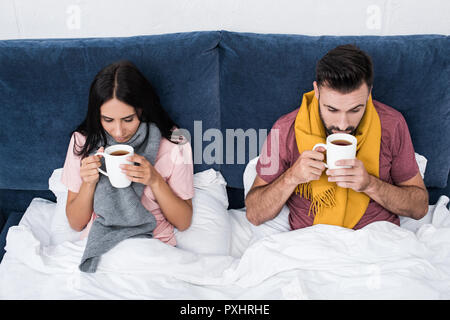 Hohe Betrachtungswinkel von kranken Jungen Paar in Schals im Bett sitzend mit Tassen heißes Getränk Stockfoto