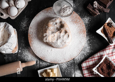 Weihnachtsplätzchen Teig auf eine hölzerne Schüssel, von Mehl, Eier, Zimt, Schokolade, Butter umgeben, und ein Nudelholz, auf einem urigen schwarz Tisch. Stockfoto