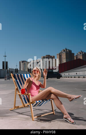 Glückliche junge Frau sitzt auf der Sonnenliege mit Cocktail in Glas auf Parkplatz Stockfoto