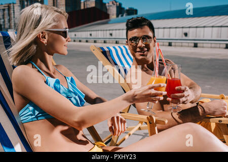 Junges Paar Anstoßen an Cocktails während der Sitzung auf Liegestühlen auf Parkplatz Stockfoto