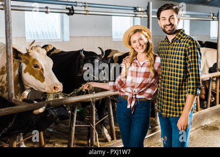 Lächelndes Paar Landwirte Fütterung von Kühen mit Heu im Stall und auf der Suche an Kamera Stockfoto