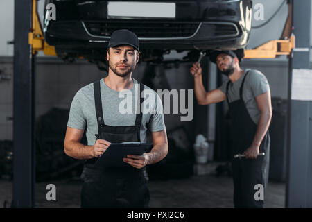 Automechaniker in Latzhosen, Zwischenablage, während Kollegen arbeiten in der Werkstatt hinter Stockfoto