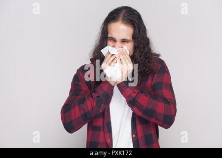 Portrait von Kälte krank stattlicher Mann mit Bart und schwarzen langen Locken im lässigen Stil, kariert rot Shirt mit Gewebe und Niesen. indoor s Stockfoto