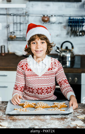 Adorable Happy Boy in santa hut hält Backblech mit Ingwer cookies und lächelnd an Kamera Stockfoto