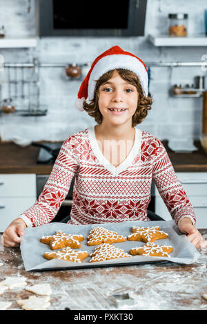 Niedliche Happy Boy in santa hut hält Backblech mit Ingwer cookies und lächelnd an Kamera Stockfoto