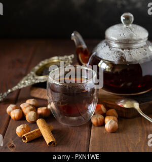 Noch immer leben Komposition. Kaffee im Glas Schale mit Teekanne in der Nähe, Vanilleschote und Haselnuss an Holz Hintergrund, mit Löffel und Sieb in der Nähe. Stockfoto