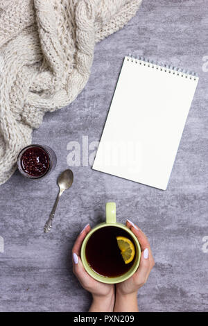 Noch immer leben und Konzept der Komposition. Tasse Tee mit Zitrone in den Händen, Strickschal, Himbeermarmelade, Löffel und Notizblock in der Nähe, am grauen Hintergrund Stockfoto