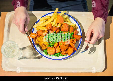 Mann mit Besteck essen und traditioneller englischer Teller - Fisch und Chips mit grünen Erbsen, gedünstete Karotte und Zahnstein Sause. Fast food, Cafe business Lun Stockfoto