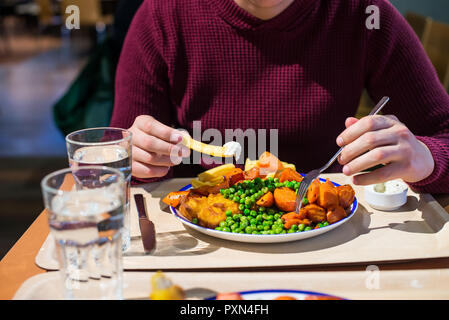 Junger Mann mit Besteck essen und traditioneller englischer Teller - Fisch und Chips mit grünen Erbsen, gedünstete Karotte und Zahnstein Sause. Fast food, Cafe busine Stockfoto