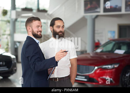 Schönen Männern stehen im Auto Center und Beobachten automobile Bärtigen. Manager smiiling, Ordner und zeigt dem Kunden Autos. Brutaler Mann im weißen Hemd weg suchen und auswählen. Stockfoto