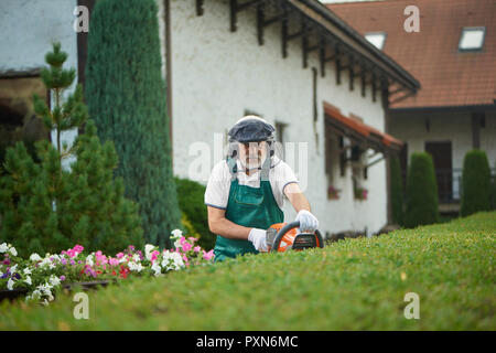 Professionelle ältere Gärtner in Uniform Schneiden von Büschen mit Scherer und Kamera. Arbeitnehmer, die in der Uniform mit Schutzmaske und schützende Kopfhörer Landschaftsgestaltung Büsche in den Hinterhof. Stockfoto