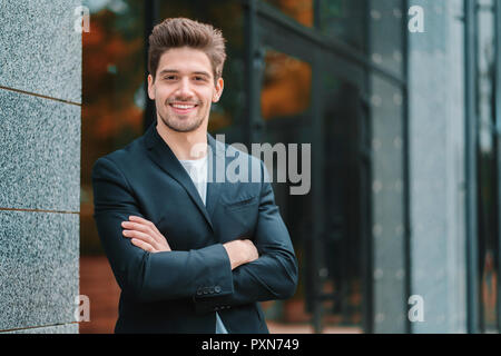 Portrait der Junge erfolgreiche zuversichtlich Geschäftsmann in der Stadt auf Bürogebäude Hintergrund. Mann im Anzug auf die Kamera schauen und lächeln. Porträts von hübscher Kerl Stockfoto