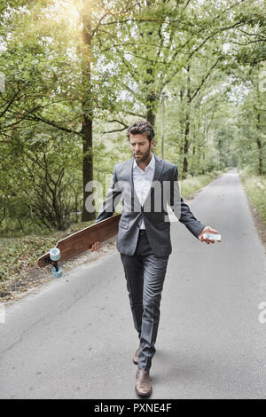 Unternehmer gehen mit Skateboard und Smartphone auf die ländliche Straße Stockfoto