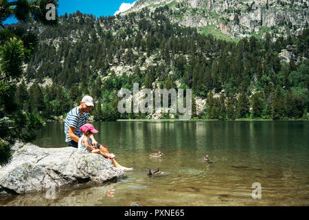 Spanien, Vater und Tochter auf einem Stein saß an einem Bergsee, feeding ducks Stockfoto